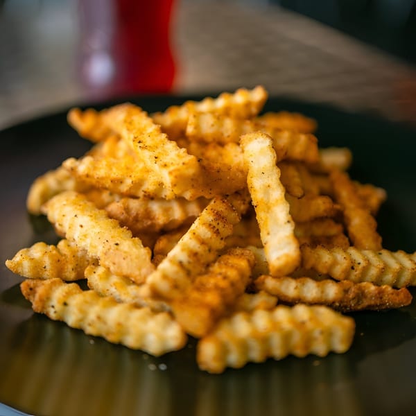 A close-up of a pile of golden, seasoned crinkle-cut fries on a dark plate.